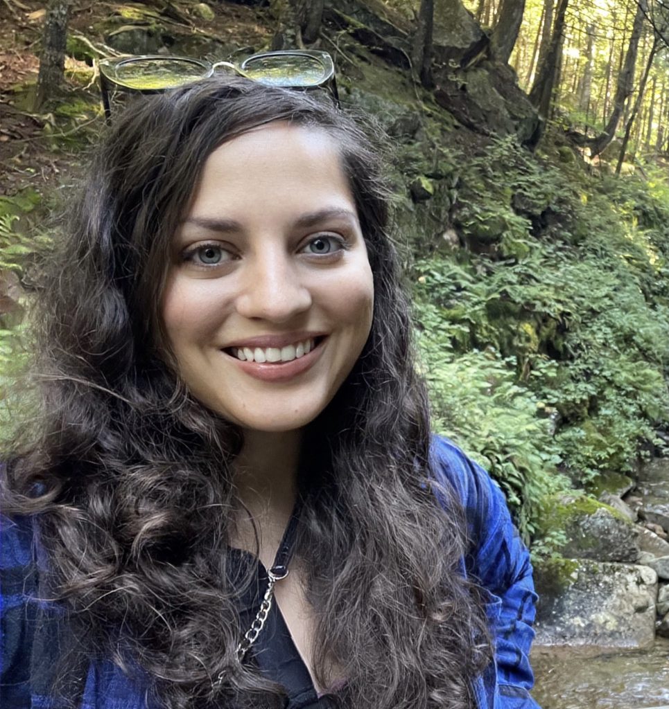 Editor Kate McCulley smiling at the camera. She has long dark curly hair and is in the middle of the woods, wearing a blue plaid shirt.