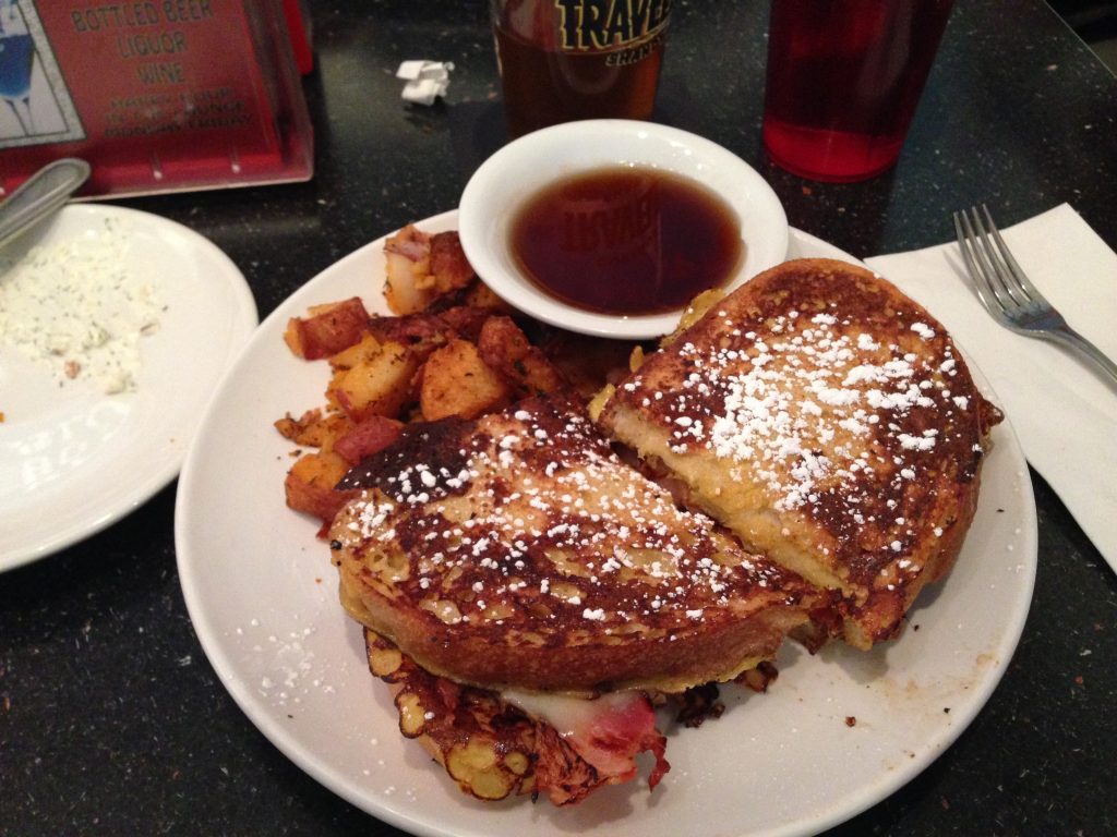 a breakfast sandwich that looks like French toast with eggs, bacon, melted cheese, next to a pile of home fries and syrup.