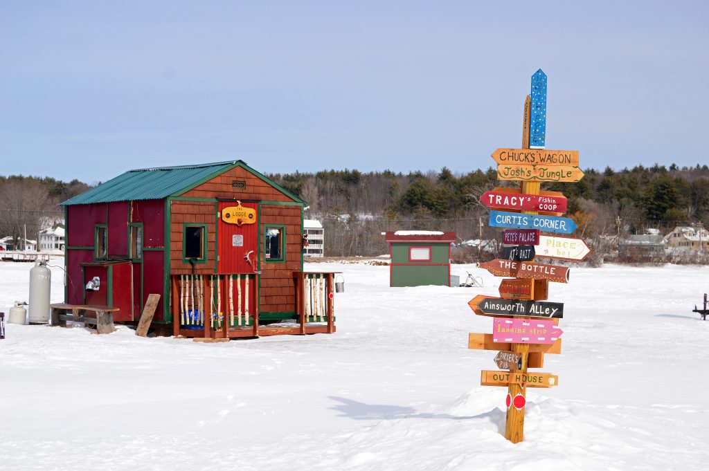 A small wooden shack on a frozen lake, next to a pole covered with wooden signs pointing to different shacks.