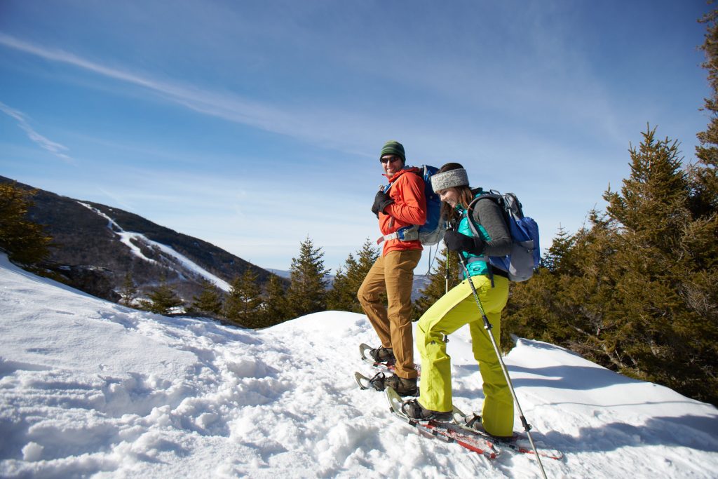 A man and a woman climbing a mountain in snowshoes, smiling underneath a blue sky.
