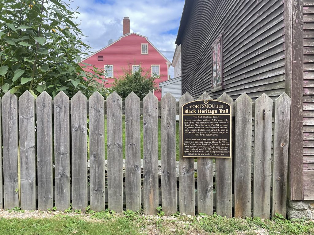 A weathered wooden fence between historic homes with a plaque reading "Portsmouth Black Heritage Trail."