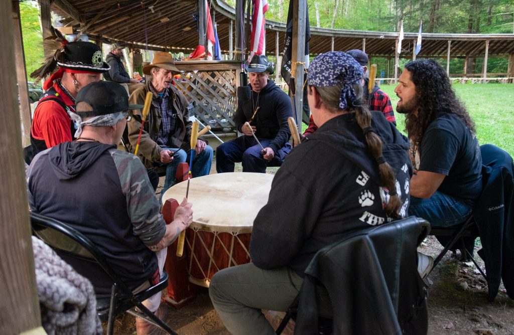 A group of Native American men, mostly dressed casually in sweatshirts and hats with some native touches, sitting in a circle and playing a drum together.