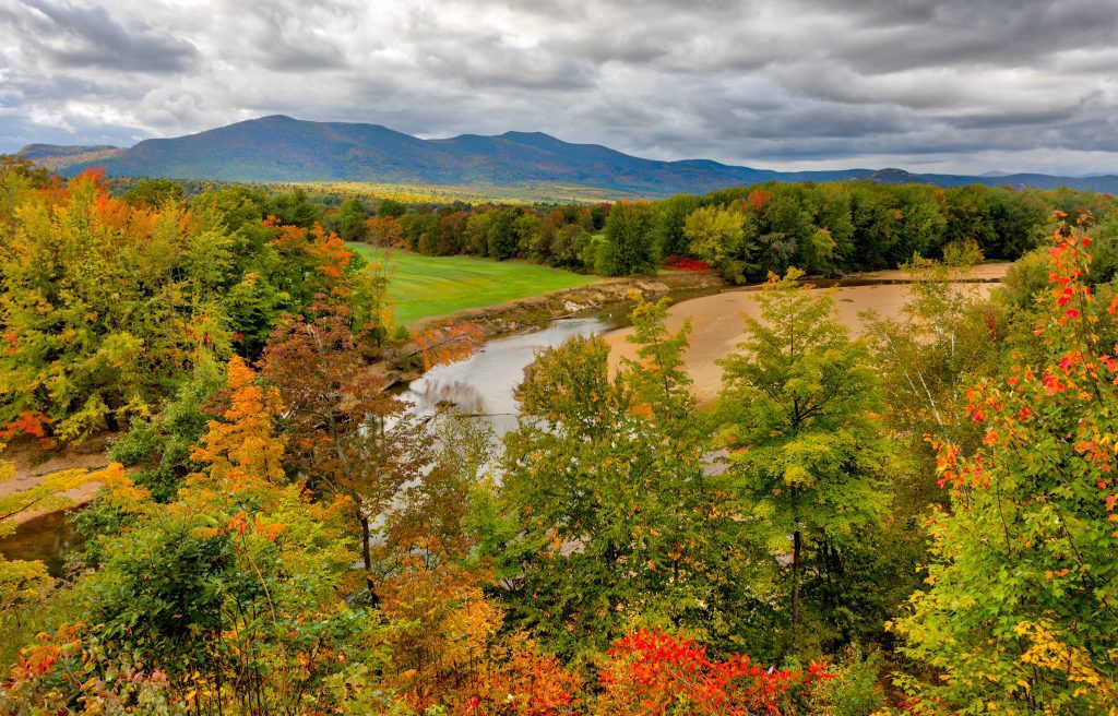 The New Hampshire countryside: mountains in the background, a river running through, and several orange, yellow, and green trees.