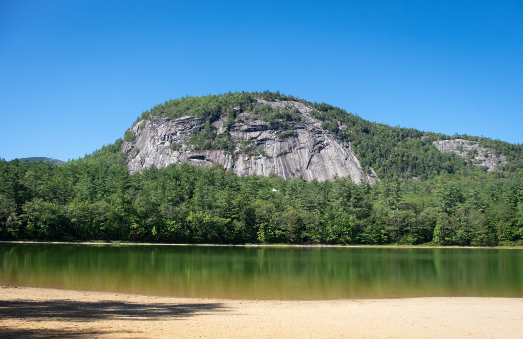 A sandy beach in front of a still green lake surrounded by pine trees, and a big gray rock face in the distance.