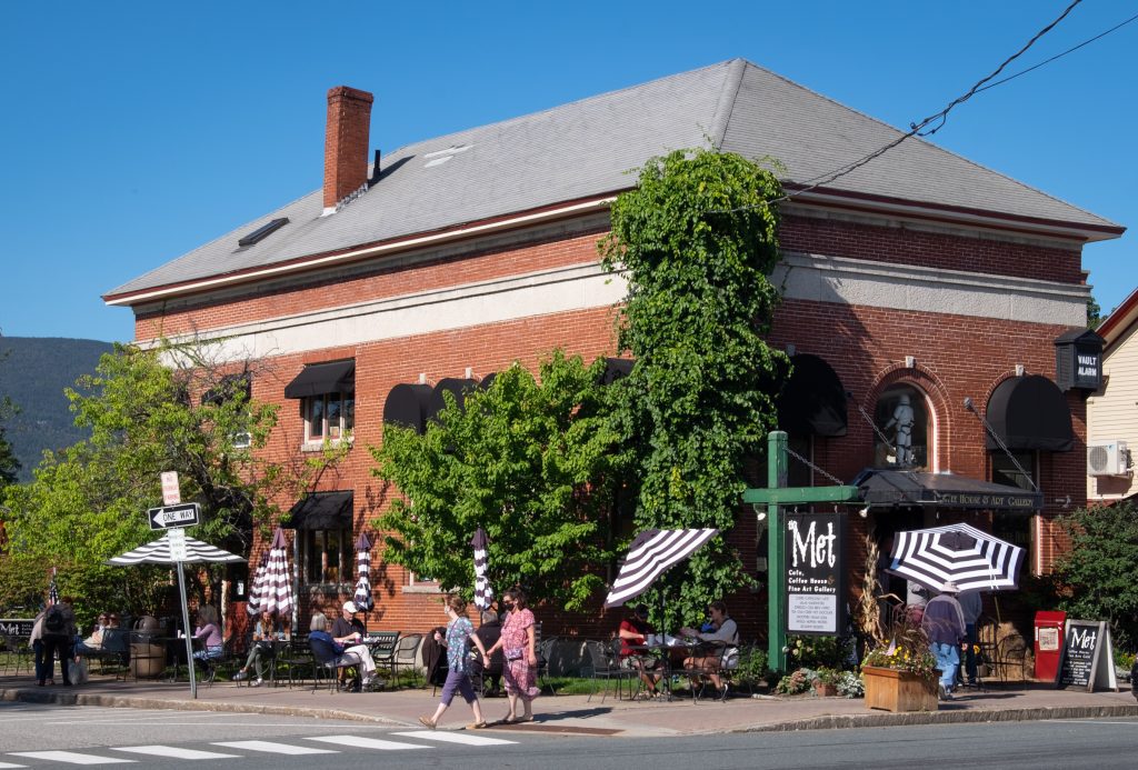 The outside of the Met Coffeehouse: a brick square building, with lots of black and white striped umbrellas outside and people dining at cafe tables.