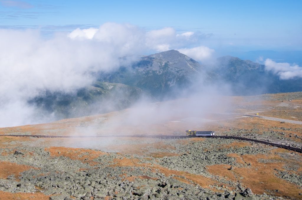 A tiny train chugging down the tracks on top of Mount Washington, a rocky landscape surrounded by cloud formations.