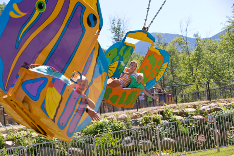 A ride with brightly colored fish swings flying through the air, parents and kids on them.