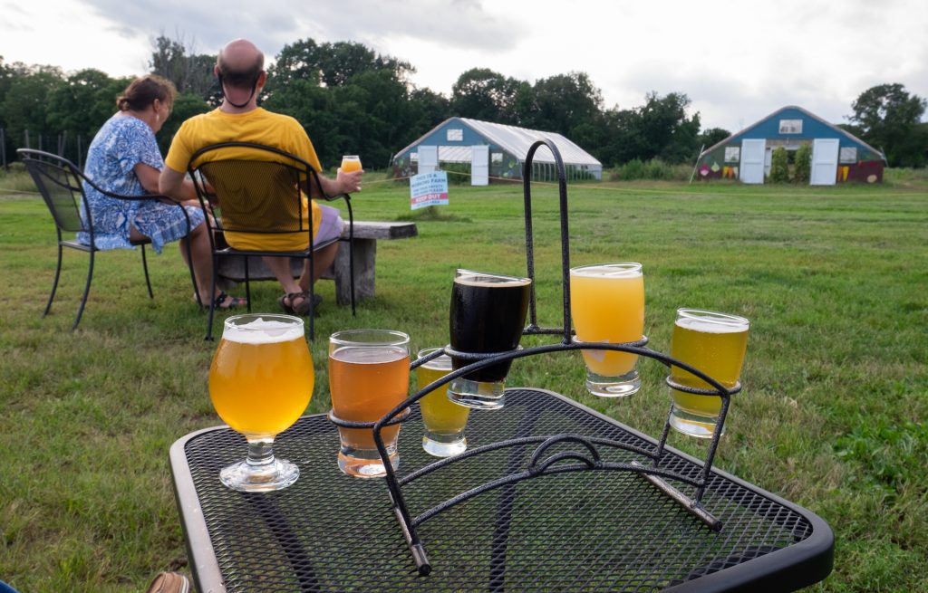 Two people sitting outdoors on the grass, with an iron table with six different beers sitting on it.
