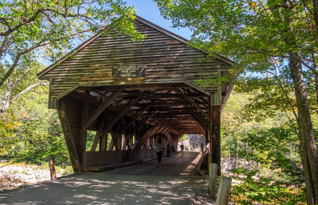 A wooden covered bridge, some people walking inside of it. You can see the beams inside it.