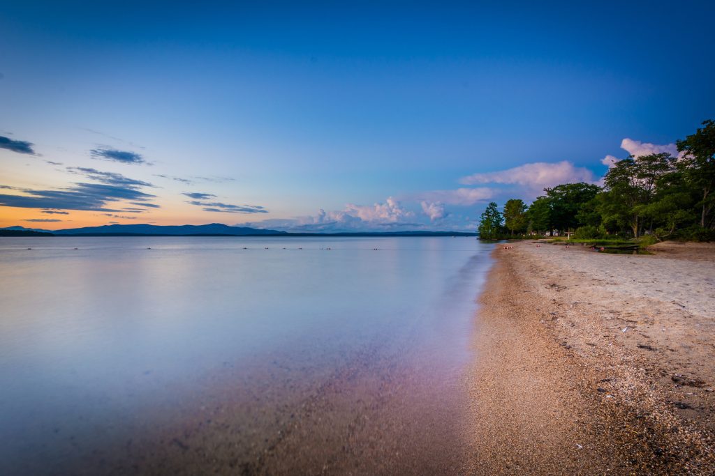 A brown sandy beach leading to a still blue lake, shot at sunset, the sky bright blue with pink clouds.
