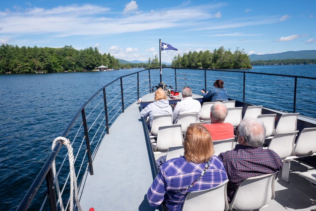 The front of a small cruise boat, several older people sitting and watching the forested islands on the lake.