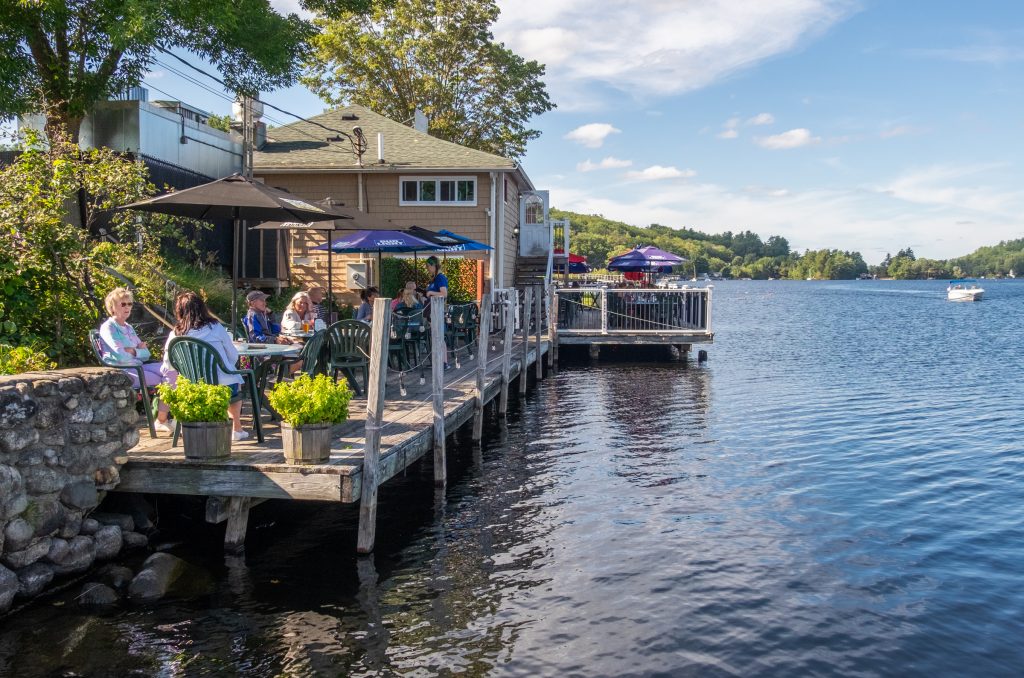 People having beers and sitting beneath umbrellas on a small wooden dock next to the lake.