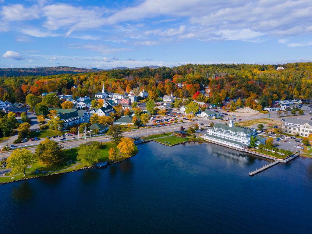 An aerial shot of the small town of Meredith NH on the edge of the bright blue lake, lots of trees in orange and yellow.
