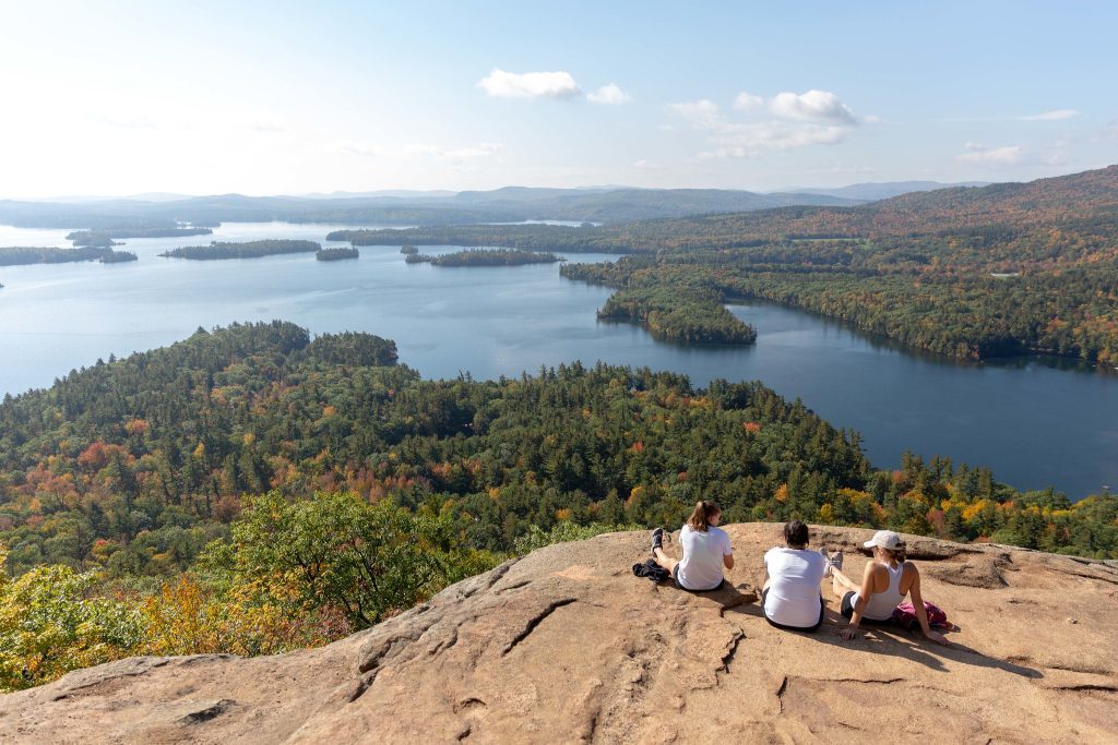 Three women hikers perched on a large rock slab looking over a view of forested islands on a calm blue lake.