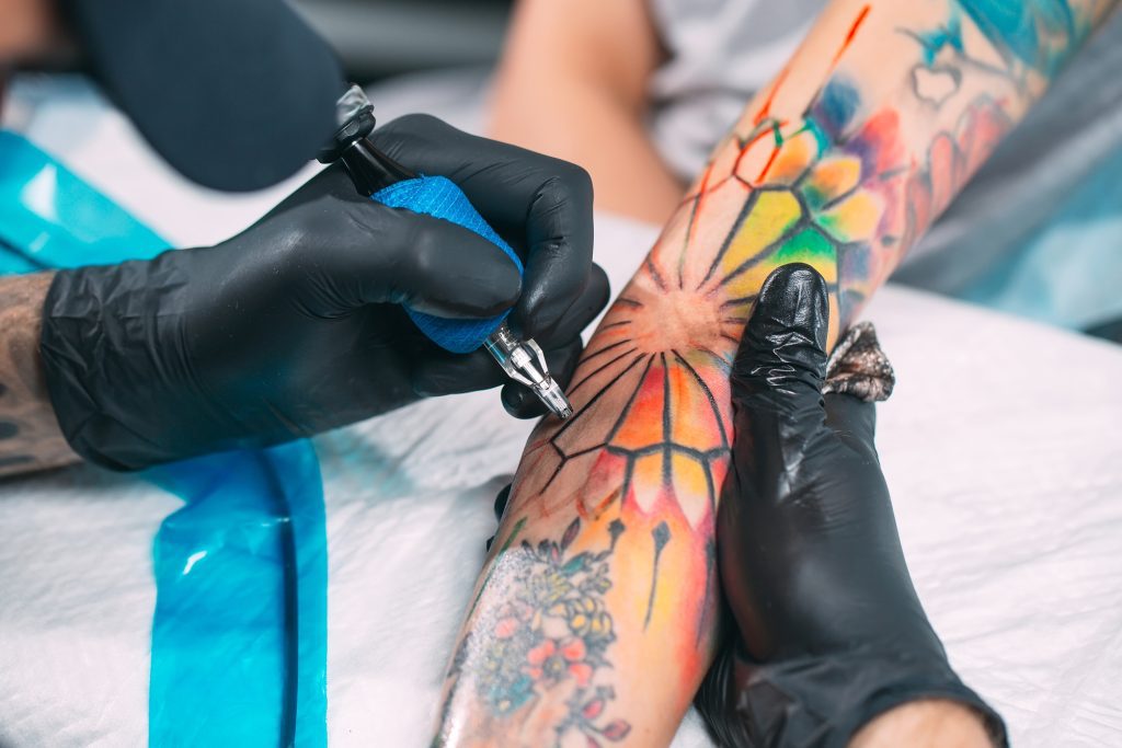 A tattoo artist wearing black gloves tattoos a rainbow design in the inside of a woman's elbow.