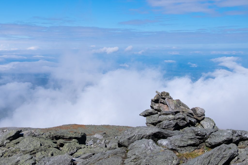 A rock cairn (pile of rocks) on the edge of the mountain. Clouds and blue sky beyond it.