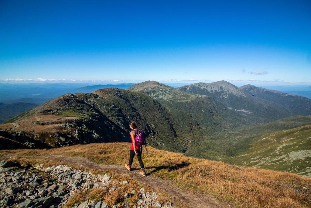A woman in shorts and a tank top hiking a bare mountain, several more mountains behind her.