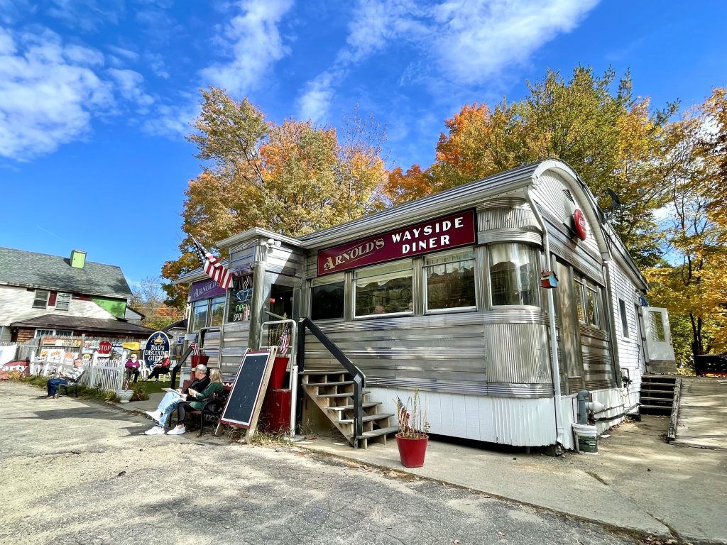 An old-fashioned diner made of shining chrome, underneath bright orange fall trees and a bright blue sky.