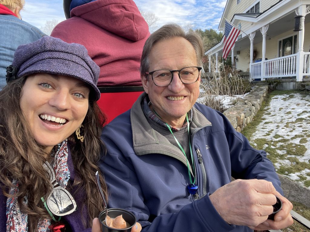 Editor Kate, in a purple hat, sitting next to her father in a sleigh, both smiling.