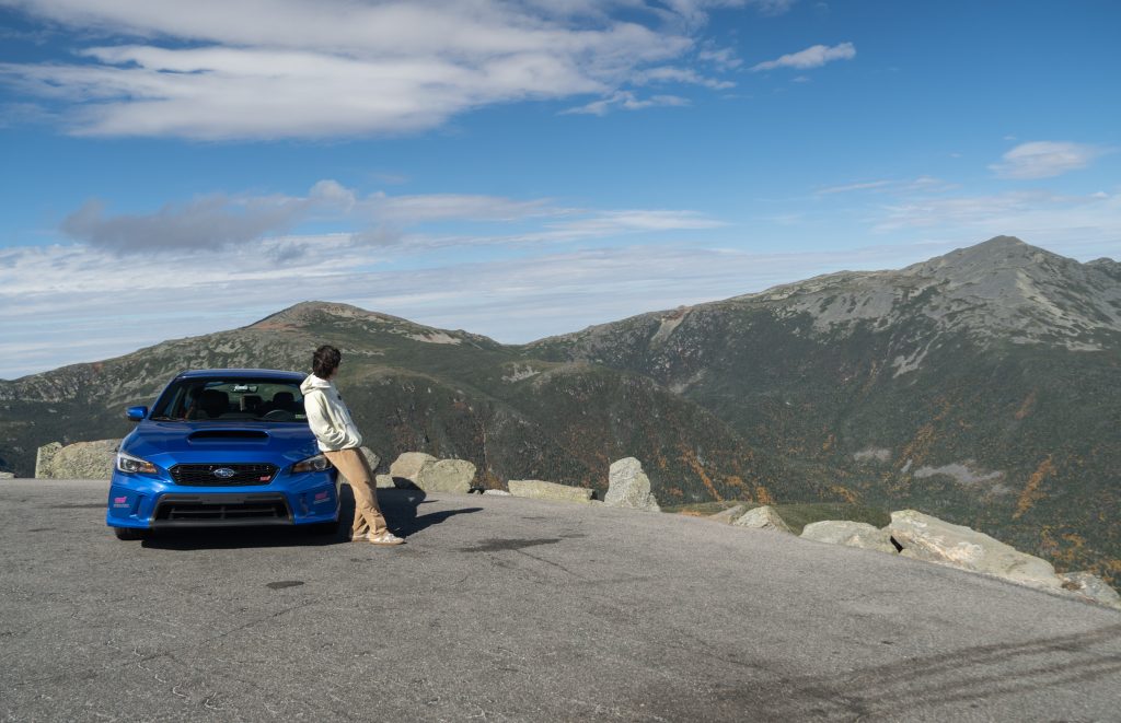 A man leaning against a sports car and looking out over the mountain range.