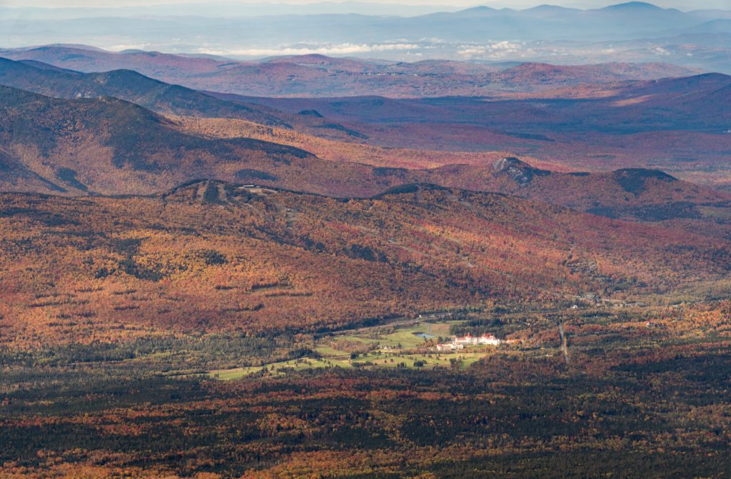A view of layers of mountains topped in bright orange foliage. You can see a resort in the distance illuminated in light.