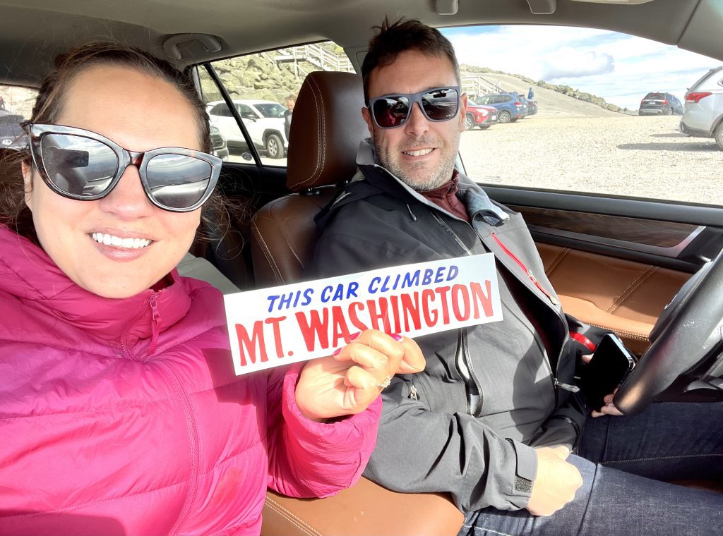 A woman in a pink jacket and a man in a black jacket take a selfie holding a "This car climbed Mt. Washington" sticker while sitting in a car.