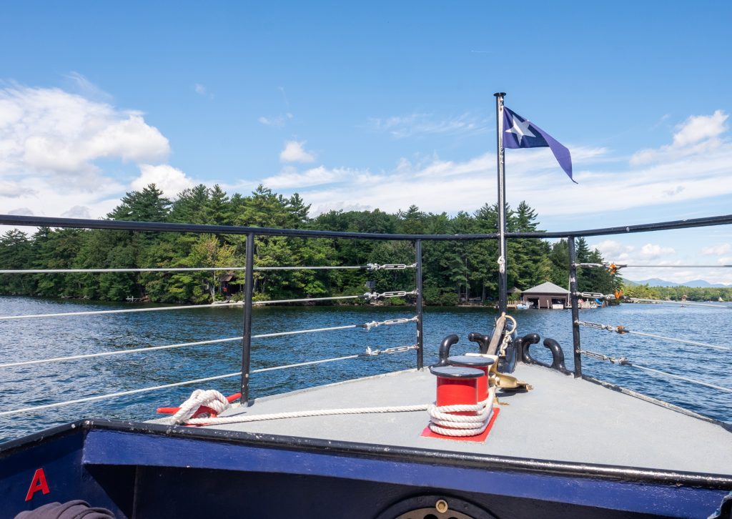 The bow of a boat sailing along a calm lake with forested islands.