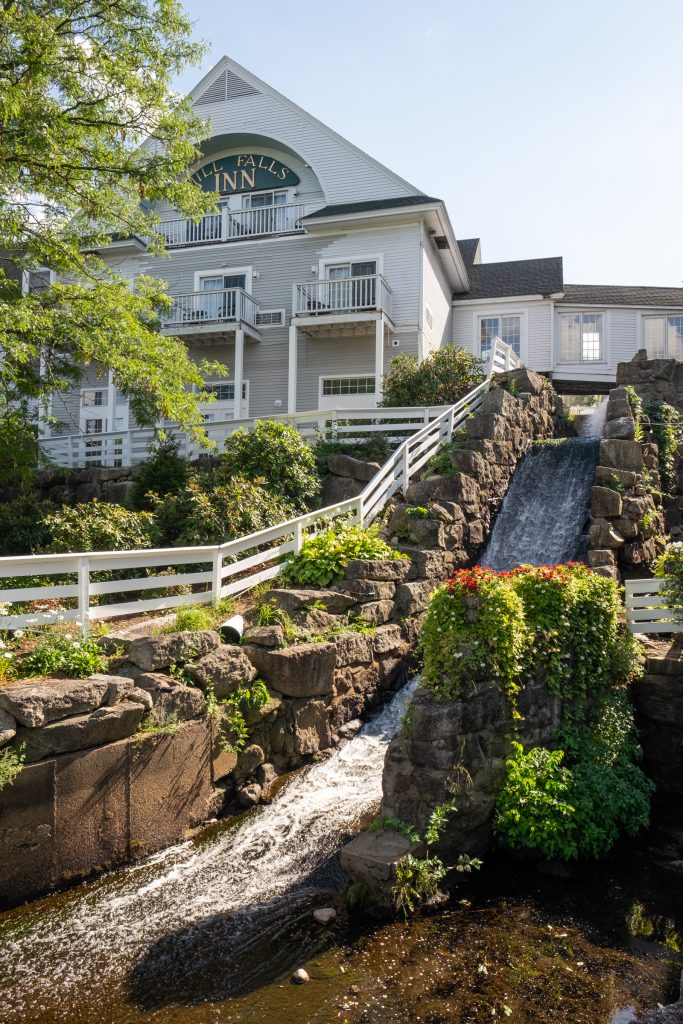 A waterfall running down a rocky hill in front of a building reading Mills Falls Marketplace.
